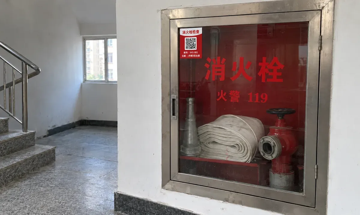 楼道里的不锈钢消火栓箱，玻璃门左上角贴着消火栓检查二维码卡片