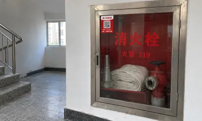 楼道里的不锈钢消火栓箱，玻璃门左上角贴着消火栓检查二维码卡片